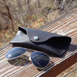 May include: Sunglasses with gold frames and dark lenses rest on a wooden surface next to a black leather case. The case has a snap closure with a silver button. The sunglasses reflect the surrounding environment.