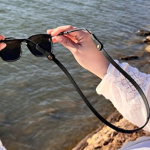 Puede incluir: Gafas de sol negras con una correa de cuero negro. Las gafas de sol tienen una montura cuadrada y lentes oscuras. La correa est&aacute; sujeta a la montura con peque&ntilde;os remaches dorados. El fondo es una masa de agua.