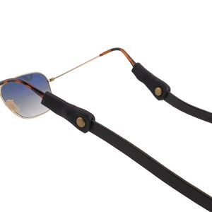 Puede incluir: Gafas de sol de aviador con lentes degradadas azules y montura dorada sujetas a una correa de gafas de cuero negro. La correa tiene extremos de goma negra con cierres de bot&oacute;n dorados.