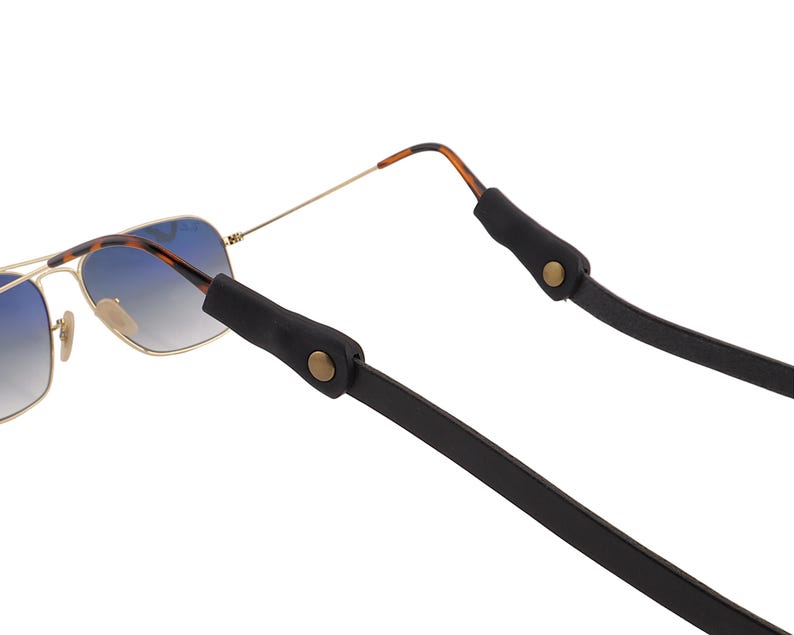 Puede incluir: Gafas de sol de aviador con lentes degradadas azules y montura dorada sujetas a una correa de gafas de cuero negro. La correa tiene extremos de goma negra con cierres de bot&oacute;n dorados.