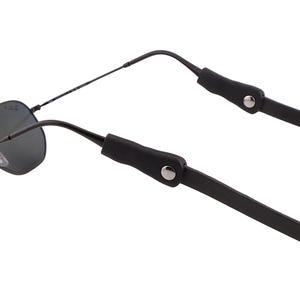 Puede incluir: Gafas de sol negras con montura met&aacute;lica y lentes oscuras. Las gafas tienen una correa de cuero negro sujeta a las patillas con remaches met&aacute;licos plateados. La correa est&aacute; dise&ntilde;ada para sujetar las gafas alrededor del cuello.