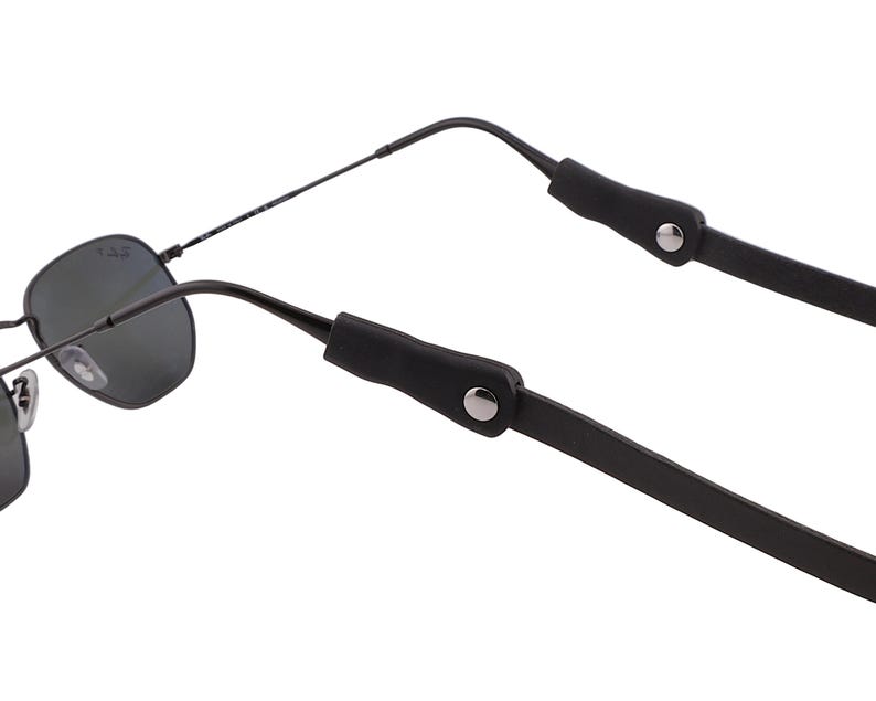 Puede incluir: Gafas de sol negras con montura met&aacute;lica y lentes oscuras. Las gafas tienen una correa de cuero negro sujeta a las patillas con remaches met&aacute;licos plateados. La correa est&aacute; dise&ntilde;ada para sujetar las gafas alrededor del cuello.