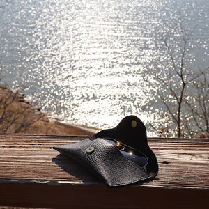 May include: Black leather sunglasses case with a snap closure, holding a pair of sunglasses. The case is open, revealing the lenses. The case is on a wooden surface with a view of a body of water in the background.