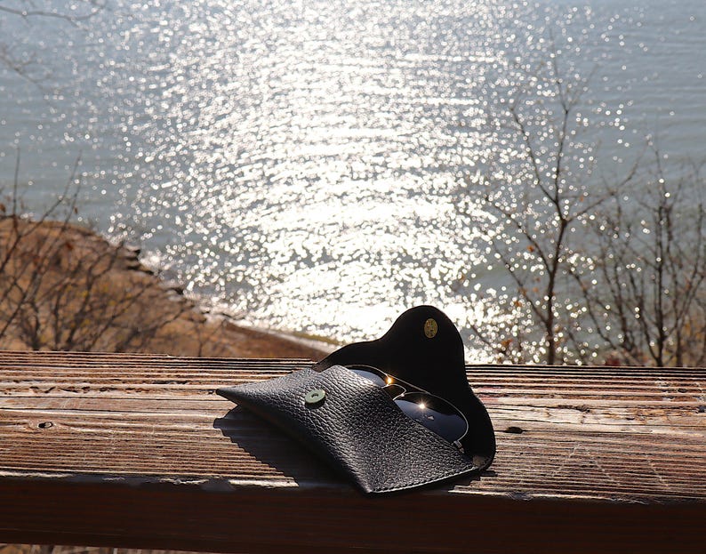 May include: Black leather sunglasses case with a snap closure, holding a pair of sunglasses. The case is open, revealing the lenses. The case is on a wooden surface with a view of a body of water in the background.