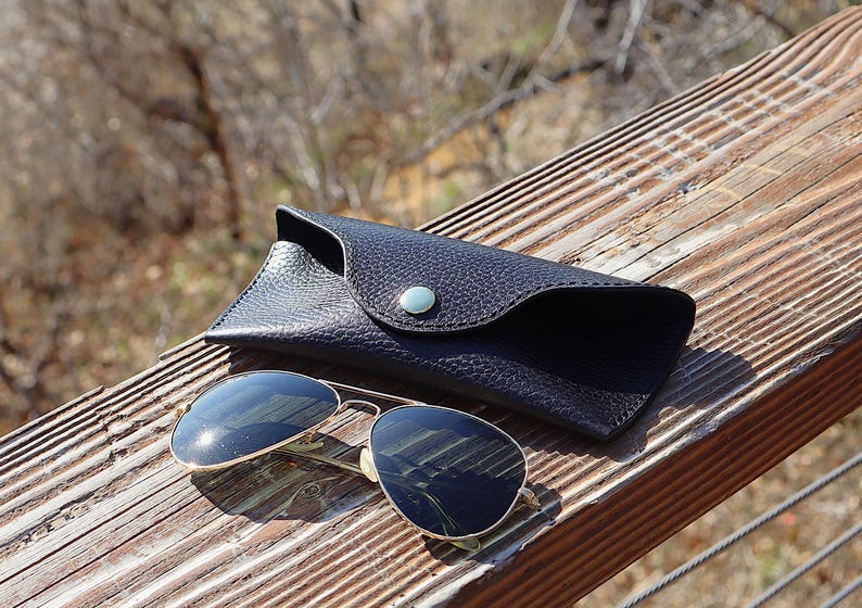 May include: Sunglasses with gold frames and dark lenses rest on a wooden surface next to a black leather case. The case has a snap closure with a silver button. The sunglasses reflect the surrounding environment.