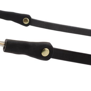 Puede incluir: Primer plano de dos correas de gafas de cuero negro con detalles met&aacute;licos dorados. Las correas tienen un dise&ntilde;o simple y minimalista, con un peque&ntilde;o bot&oacute;n dorado en cada correa. Las puntas de las correas son doradas.