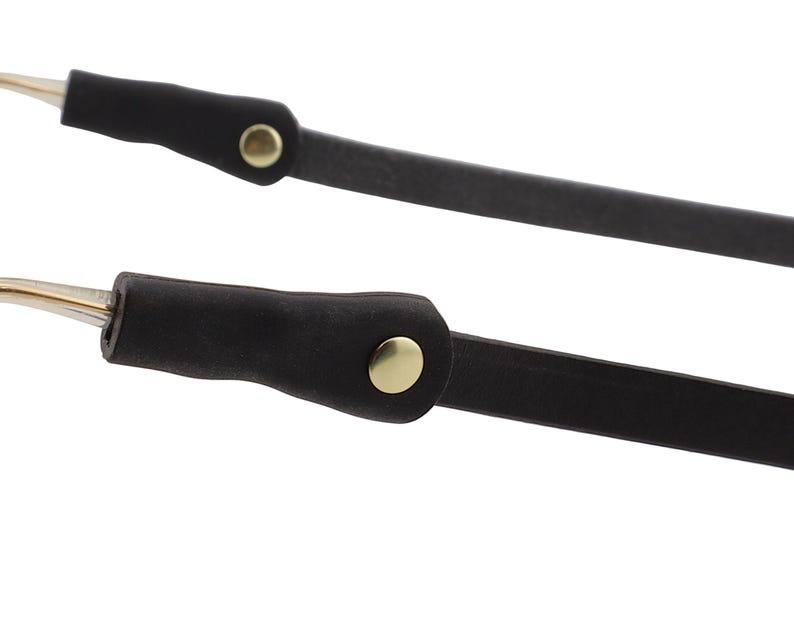 Puede incluir: Primer plano de dos correas de gafas de cuero negro con detalles met&aacute;licos dorados. Las correas tienen un dise&ntilde;o simple y minimalista, con un peque&ntilde;o bot&oacute;n dorado en cada correa. Las puntas de las correas son doradas.