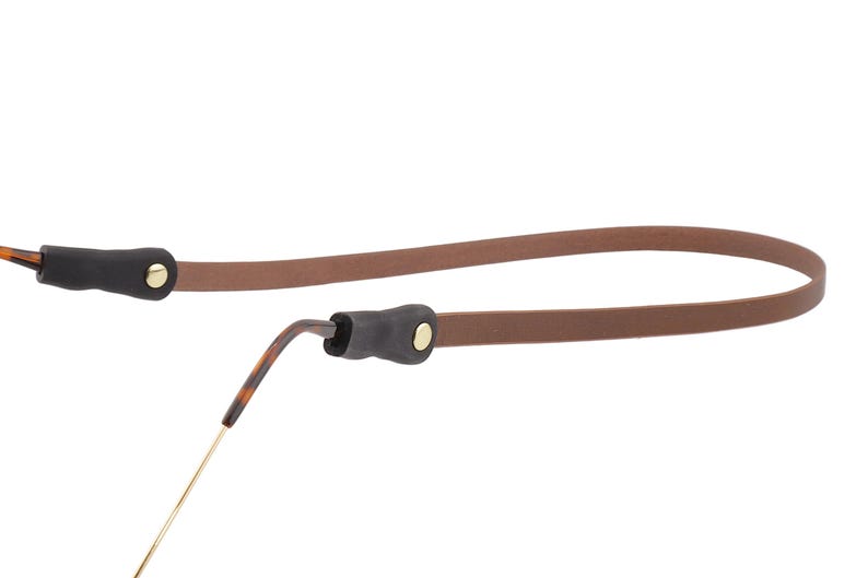 Puede incluir: Una correa de gafas de cuero marr&oacute;n con extremos de goma negra y remaches dorados. La correa est&aacute; dise&ntilde;ada para sujetar las gafas y evitar que se caigan. Las gafas tienen un patr&oacute;n de carey.