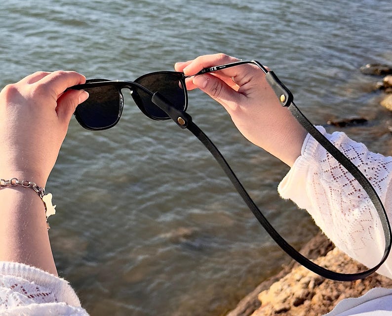 Puede incluir: Gafas de sol negras con una correa de cuero negro. Las gafas de sol tienen una montura cuadrada y lentes oscuras. La correa est&aacute; sujeta a la montura con peque&ntilde;os remaches dorados. El fondo es una masa de agua.