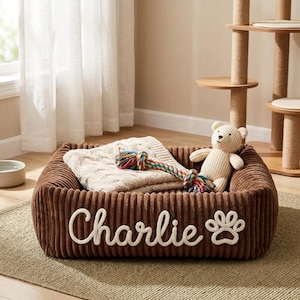 以下が含まれることがあります： 茶色のコーデュロイの犬用ベッド。白い筆記体で「Charlie」という名前と足跡が描かれています。ベッドにはクリーム色の毛布、ロープのおもちゃ、テディベアが入っています。