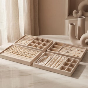 Puede incluir: Organizadores de joyas de terciopelo beige llenos de anillos, collares, pulseras y pendientes. Los organizadores están colocados sobre una mesa con tapa de mármol, con jarrones de tonos neutros en el fondo. Las joyas son doradas y de perlas.