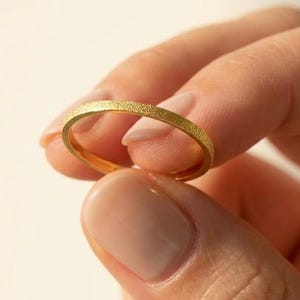 May include: A gold ring with a textured surface is held between fingers. The ring has a simple band design and a matte finish. The ring appears to be a wedding band or a minimalist style ring.