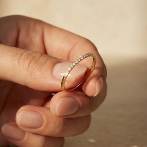 Könnte beinhalten: Ein zarter Goldring mit einer Reihe kleiner, funkelnder Edelsteine. Der Ring wird in einer Hand gehalten, wodurch das schlanke Band und das subtile Funkeln der Steine hervorgehoben werden. Das Design des Rings ist schlicht und elegant.