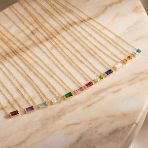Pode incluir: Uma coleção de colares de ouro com pedras preciosas retangulares em várias cores, incluindo vermelho, roxo, azul e verde. Os colares são exibidos em uma superfície de mármore, mostrando suas delicadas correntes e pedras vibrantes. As pedras são definidas em biséis de ouro.
