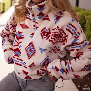 Puede incluir: Un jersey de forro polar blanco con cuello alto y cremallera de un cuarto. El jersey presenta un estampado de formas geométricas rojas y azules. Las mangas tienen puños elásticos. El material de forro polar parece suave y cálido.