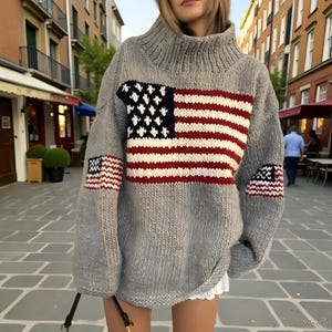 May include: A grey, oversized turtleneck jumper with a large American flag design on the front and smaller flags on the sleeves. The flag design features red and white stripes with a blue field of white stars.
