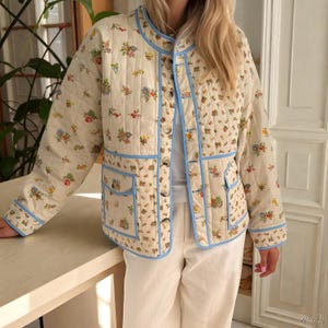 May include: Cream-colored quilted jacket with a floral pattern in red, blue, and green. The jacket features a light blue trim, button closures, and two front pockets. The jacket is a lightweight outerwear garment.