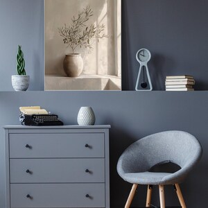 Op de afbeelding: Een serene interieursc&egrave;ne met een grijze dressoir, een grijze stoel met houten poten en een schilderij van een vaas met olijftakken. Een kleine klok en boeken staan op een plank erboven.