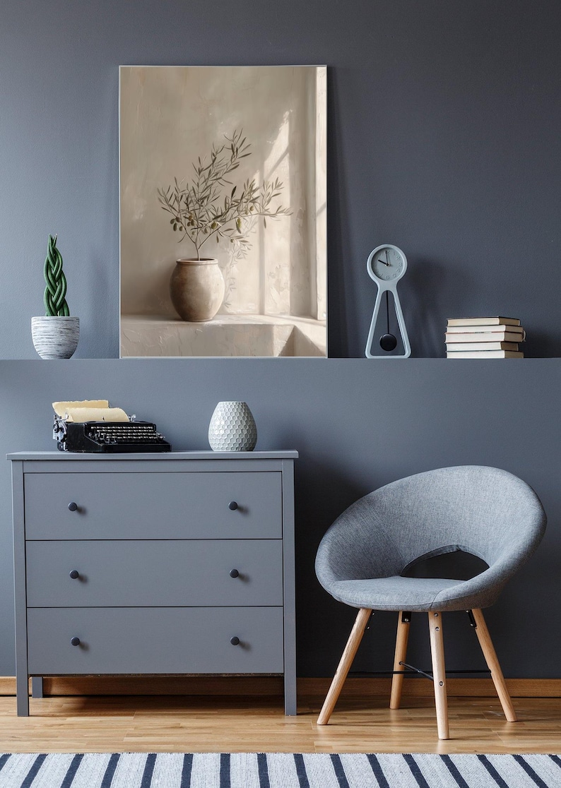 Op de afbeelding: Een serene interieursc&egrave;ne met een grijze dressoir, een grijze stoel met houten poten en een schilderij van een vaas met olijftakken. Een kleine klok en boeken staan op een plank erboven.