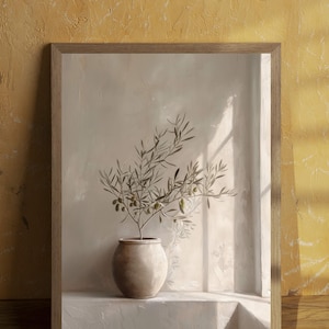Op de afbeelding: Een ingelijste print met een olijftak in een beige keramische pot. Het kunstwerk staat tegen een neutraal getinte muur met een raam dat schaduwen werpt. De lijst is van lichtgekleurd hout en de algehele esthetiek is minimalistisch.