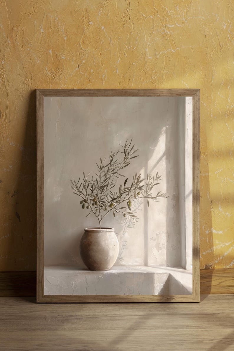 Op de afbeelding: Een ingelijste print met een olijftak in een beige keramische pot. Het kunstwerk staat tegen een neutraal getinte muur met een raam dat schaduwen werpt. De lijst is van lichtgekleurd hout en de algehele esthetiek is minimalistisch.