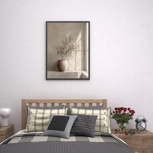 Op de afbeelding: Een ingelijste prent met een stilleven van een vaas met olijftakken. Het kunstwerk hangt boven een bed met geruite kussens en een grijze sprei. Een houten nachtkastje bevat boeken, een witte vaas en een kleine wekker.