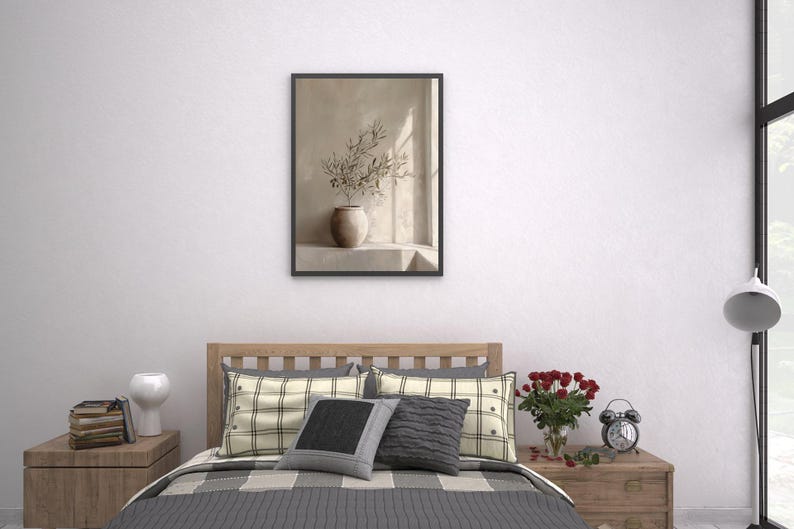 Op de afbeelding: Een ingelijste prent met een stilleven van een vaas met olijftakken. Het kunstwerk hangt boven een bed met geruite kussens en een grijze sprei. Een houten nachtkastje bevat boeken, een witte vaas en een kleine wekker.
