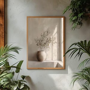 Op de afbeelding: Een ingelijste print met een olijftak in een beige keramische vaas. Het kunstwerk staat tegen een neutrale achtergrond met zachte verlichting, wat een serene en minimalistische esthetiek cre&euml;ert. De lijst is lichtbruin.