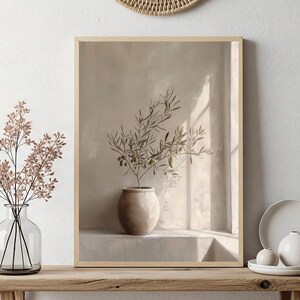 Op de afbeelding: Een ingelijste prent van een olijftak in een beige pot, tegen een neutrale achtergrond. Een heldere glazen vaas met roze takken en een kleine witte vaas met een bord en twee bollen staan op een houten plank.