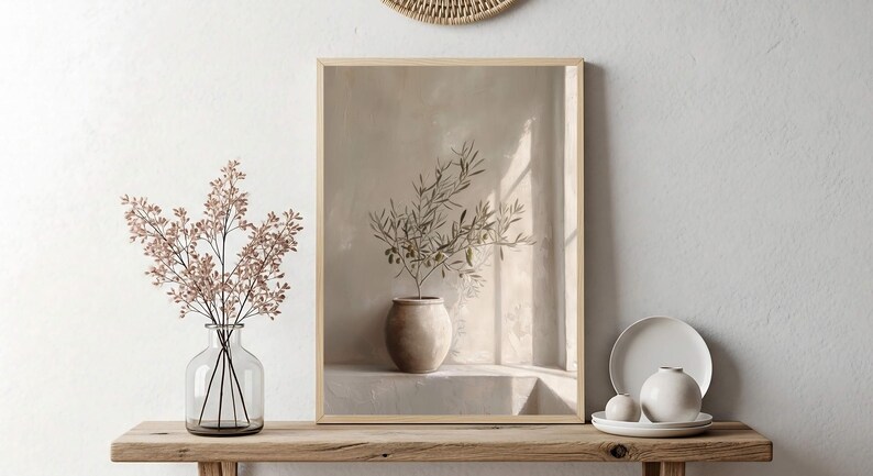 Op de afbeelding: Een ingelijste prent van een olijftak in een beige pot, tegen een neutrale achtergrond. Een heldere glazen vaas met roze takken en een kleine witte vaas met een bord en twee bollen staan op een houten plank.