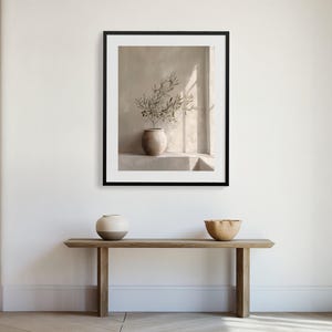 Op de afbeelding: Een ingelijste prent met een stilleven van een vaas met olijftakken tegen een neutrale achtergrond. Daaronder een houten console met een keramische vaas en een houten kom. De sc&egrave;ne wordt badend in natuurlijk licht.
