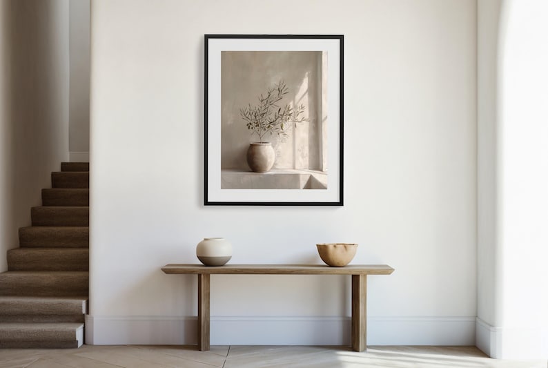 Op de afbeelding: Een ingelijste prent met een stilleven van een vaas met olijftakken tegen een neutrale achtergrond. Daaronder een houten console met een keramische vaas en een houten kom. De sc&egrave;ne wordt badend in natuurlijk licht.