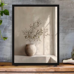Op de afbeelding: Een ingelijste kunstdruk met een stilleven. Het toont een olijftak in een keramische pot, tegen een neutraal getinte muur met zonlicht dat door een raam schijnt. De lijst is zwart.