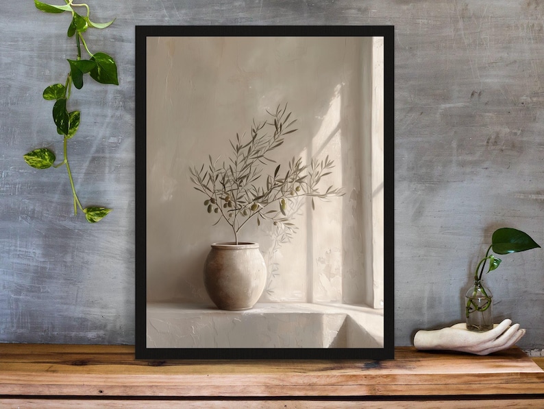 Op de afbeelding: Een ingelijste kunstdruk met een stilleven. Het toont een olijftak in een keramische pot, tegen een neutraal getinte muur met zonlicht dat door een raam schijnt. De lijst is zwart.