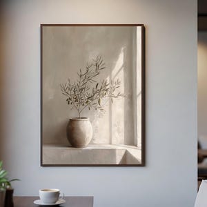 Op de afbeelding: Een ingelijst kunstwerk toont een beige vaas met olijftakken. Het kunstwerk staat tegen een neutrale achtergrond, met zonlicht dat schaduwen cre&euml;ert. Het kunstwerk wordt aan een witte muur getoond. De afbeelding is in een caf&eacute;.