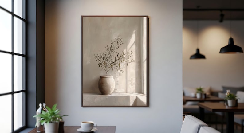 Op de afbeelding: Een ingelijst kunstwerk toont een beige vaas met olijftakken. Het kunstwerk staat tegen een neutrale achtergrond, met zonlicht dat schaduwen cre&euml;ert. Het kunstwerk wordt aan een witte muur getoond. De afbeelding is in een caf&eacute;.