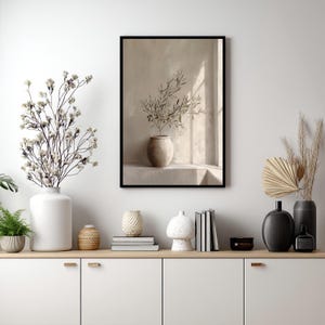 Op de afbeelding: Een ingelijste print met een beige vaas met olijftakken, tegen een neutrale muur met een raam. De afbeelding staat in een zwarte lijst. De sc&egrave;ne maakt deel uit van een grotere display van decoratieve items op een witte kast.