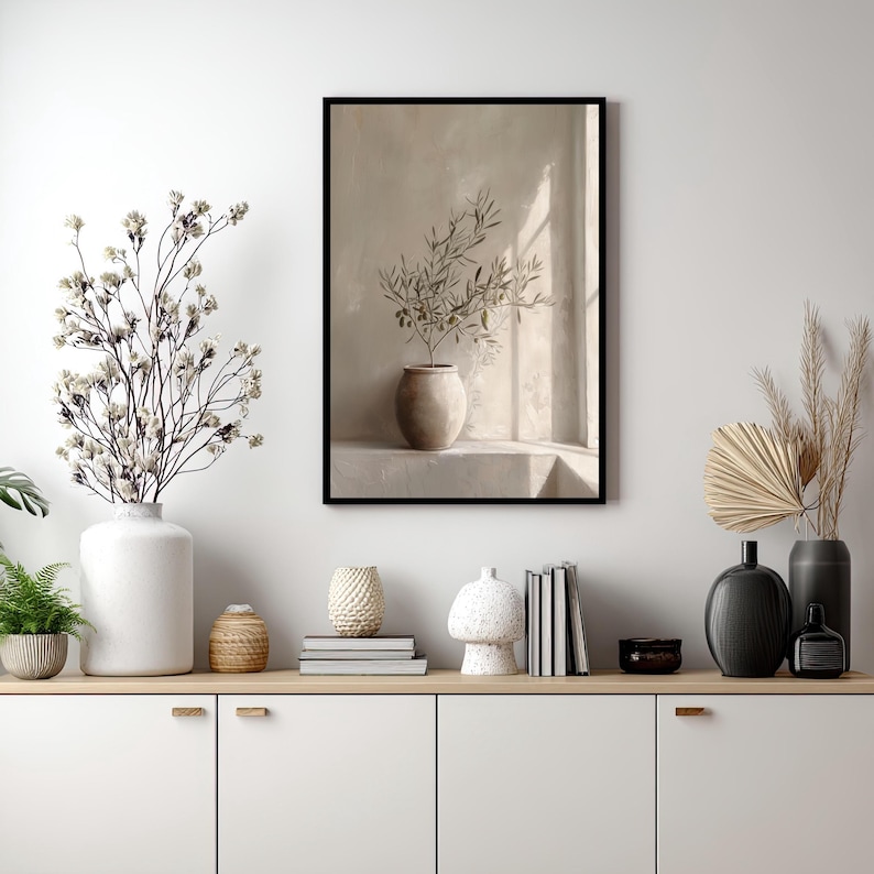 Op de afbeelding: Een ingelijste print met een beige vaas met olijftakken, tegen een neutrale muur met een raam. De afbeelding staat in een zwarte lijst. De sc&egrave;ne maakt deel uit van een grotere display van decoratieve items op een witte kast.