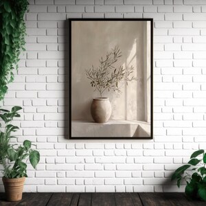 Op de afbeelding: Een ingelijste kunstdruk met olijftakken in een beige vaas. Het kunstwerk staat tegen een neutrale achtergrond met zachte verlichting. De print wordt weergegeven op een witte bakstenen muur, met planten in potten op de voorgrond.