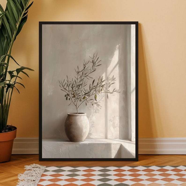 Op de afbeelding: Een ingelijste kunstdruk met een olijftak in een beige keramische vaas. Het kunstwerk staat tegen een neutrale muur met zonlicht dat schaduwen cre&euml;ert. De lijst is zwart.
