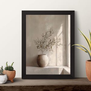 Op de afbeelding: Een ingelijste print met een olijftak in een keramische vaas. De print heeft een beige tint en zit in een zwarte lijst. Verschillende potplanten staan op een houten oppervlak.