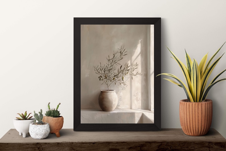 Op de afbeelding: Een ingelijste print met een olijftak in een keramische vaas. De print heeft een beige tint en zit in een zwarte lijst. Verschillende potplanten staan op een houten oppervlak.
