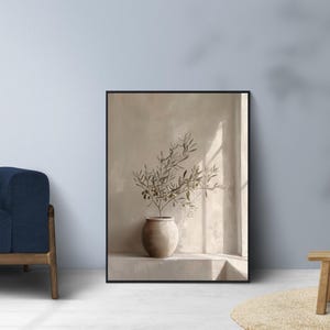 Op de afbeelding: Een ingelijste kunstdruk met een beige vaas met olijftakken. De vaas staat op een richel tegen een neutrale muur, met zonlicht dat door een raam schijnt. Het kunstwerk is in een zwart frame.