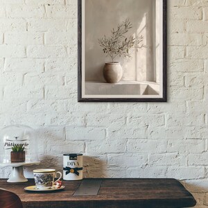 Op de afbeelding: Een ingelijste prent toont een stilleven met een beige vaas met olijftakken. Het kunstwerk staat tegen een neutrale achtergrond, met licht en schaduw. De lijst is donkerbruin.