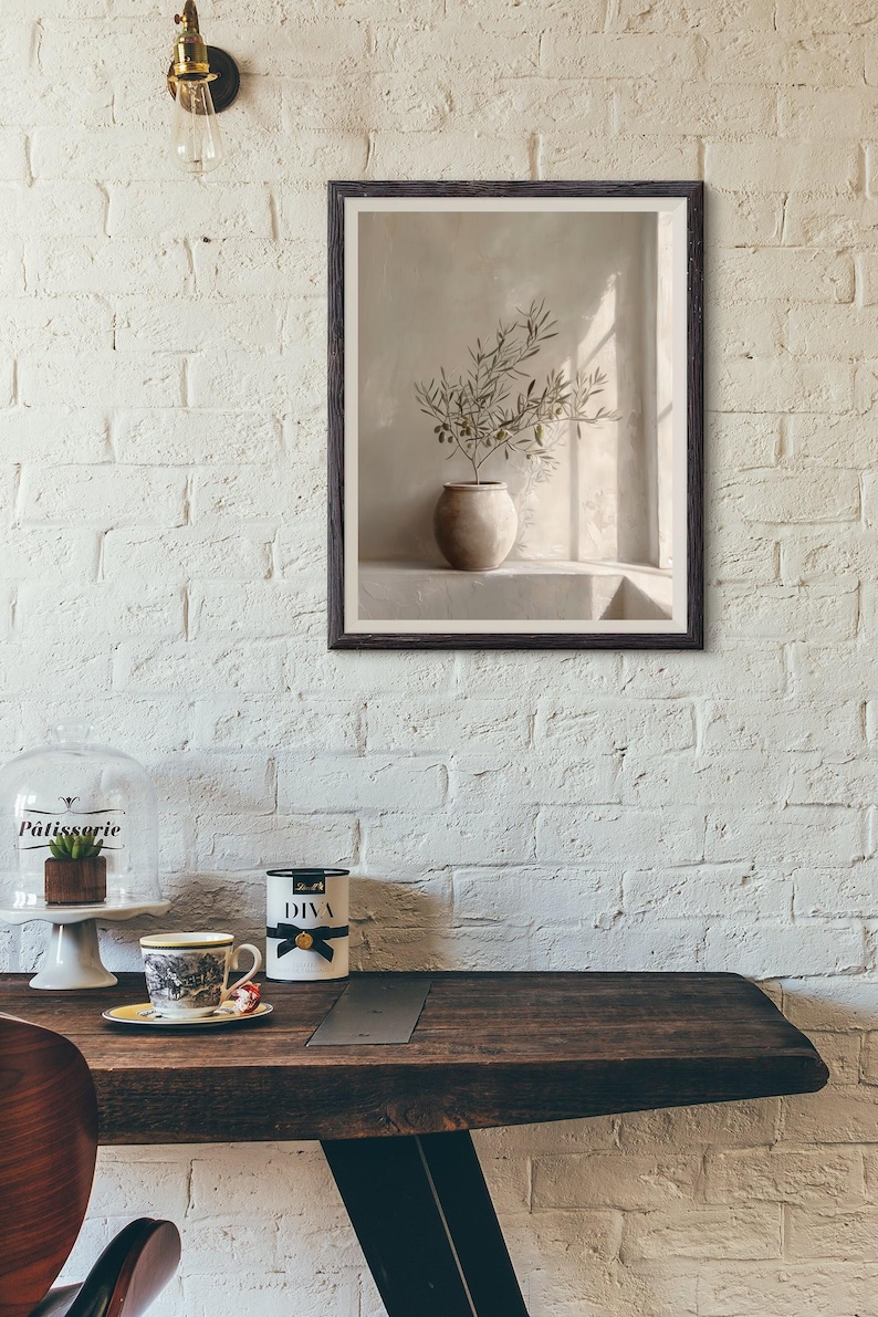 Op de afbeelding: Een ingelijste prent toont een stilleven met een beige vaas met olijftakken. Het kunstwerk staat tegen een neutrale achtergrond, met licht en schaduw. De lijst is donkerbruin.