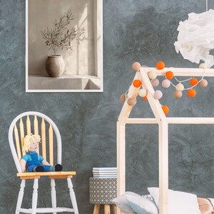 Op de afbeelding: Een ingelijste prent van een stilleven met een vaas met olijftakken. De afbeelding wordt weergegeven op een grijze muur met textuur. Een houten bedframe in de vorm van een huis is versierd met lichtslingers. Een pop zit op een wit-gele stoel.
