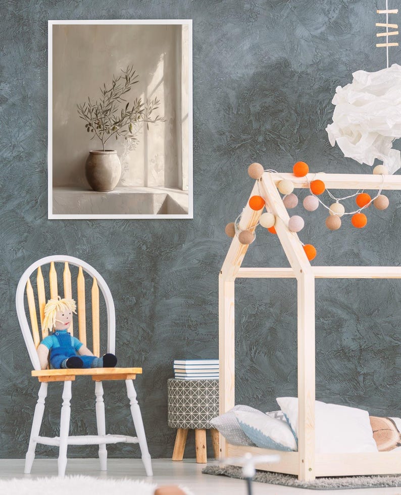Op de afbeelding: Een ingelijste prent van een stilleven met een vaas met olijftakken. De afbeelding wordt weergegeven op een grijze muur met textuur. Een houten bedframe in de vorm van een huis is versierd met lichtslingers. Een pop zit op een wit-gele stoel.