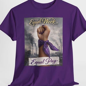 Puede incluir: Camiseta morada con un puño en alto, un pañuelo morado y las palabras "Equal Work" y "Equal Pay" en escritura dorada. El fondo muestra chimeneas industriales. Una camiseta reivindicativa para la justicia social.