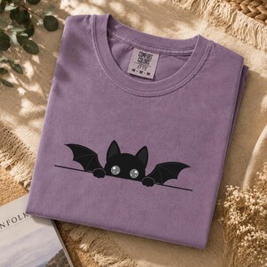 Puede incluir: Una camiseta de manga corta de color morado con un gráfico de un murciélago negro con grandes ojos blancos asomándose sobre una línea horizontal. La etiqueta interior del cuello muestra el texto Comfort Colors 1717 M M M. Tela de color sólido.