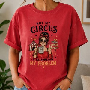 Puede incluir: Una camiseta roja de manga corta con un gráfico de una persona con diadema roja y gafas de sol sosteniendo una taza. El texto dice NOT MY CIRCUS, STILL SOMEHOW MY PROBLEM, y la taza dice NOT MY CIRCUS NOT MY MONKEYS, con una carpa de circo.